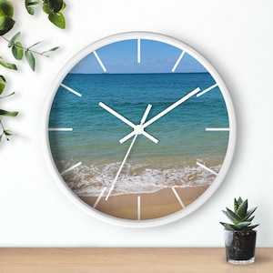 May include: White clock with a beach scene on the face. The scene shows blue ocean water with white waves crashing on a sandy beach.