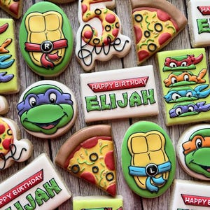 May include: A collection of decorated sugar cookies featuring Teenage Mutant Ninja Turtles and pizza slices. Some cookies are shaped like the turtles, others have the number "5", and some say "Happy Birthday Elijah". The cookies are on a wooden surface.