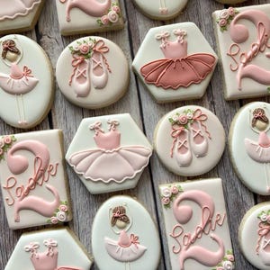 May include: An assortment of decorated sugar cookies with a ballet theme. Cookies are in various shapes, including oval, round, and rectangular, and are decorated with pink ballet shoes, tutus, and a ballerina. Some cookies have the name "Sadie" and the number "2".