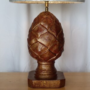 May include: A gold and red table lamp with a pine cone shaped base. The lamp has a white fabric shade.