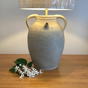 May include: A table lamp featuring a beige, textured ceramic jug base with two side handles and a small spout. The lamp is topped with a cylindrical, light-coloured fabric lampshade. A cluster of small white flowers sits at the base of the lamp.