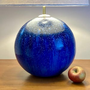 May include: A blue ceramic table lamp with a round base and a grey lampshade. The lamp has a speckled pattern and a brass neck. A red and yellow apple sits on the wooden surface.