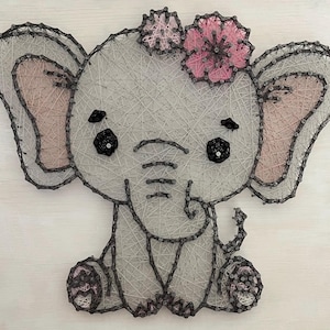 May include: A string art elephant with a pink flower and bow. The elephant is gray with pink accents on the ears and feet. The eyes are black, and the elephant is sitting down. The art is made with nails and string.