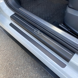 Könnte beinhalten: Schwarze Autotürschwelle mit silberner Verkleidung und weißem Chevrolet-Logo. Die Schwelle hat zwei schwarze Streifen, die über die gesamte Länge der Schwelle verlaufen.