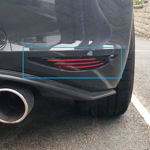 May include: A close-up of a black car's rear bumper with a red and black tail light. The tail light is a horizontal, rectangular shape with a black housing and a red lens. The tail light is located on the right side of the bumper.