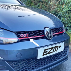 Peut inclure: Une Volkswagen Golf GTI grise avec une garniture rouge sur la calandre avant. La calandre a un motif en nid d'abeille noir et le logo GTI est en rouge. La voiture a une plaque d'immatriculation noire avec le texte "EZM+" en blanc.