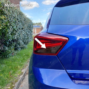 Puede incluir: Un coche azul con una luz trasera tintada. La luz trasera está parcialmente cubierta con un tinte de humo oscuro. El coche está aparcado en una calle con hierba verde y un seto en el fondo.