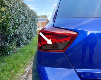 May include: A blue car with a tinted taillight. The taillight is partially covered with a dark smoke tint. The car is parked on a street with green grass and a hedge in the background.