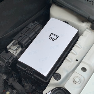 Puede incluir: Una caja de plástico blanca con una etiqueta negra que dice "EZM" y un símbolo de un enchufe. La caja está en el compartimento del motor de un coche.