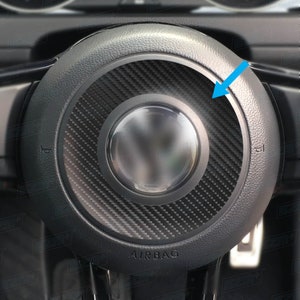 May include: Close-up of a black steering wheel with a carbon fibre trim and the word "AIRBAG" printed on the bottom. The trim is a thin ring around the centre of the steering wheel.