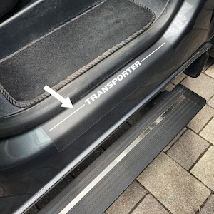 Könnte beinhalten: Nahaufnahme einer grauen Autotür mit einem weißen "TRANSPORTER"-Aufkleber an der Seite. Das Auto hat eine schwarze Trittleiste mit einem silbernen Metallstreifen.