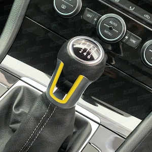 May include: A black leather gear shift knob with a yellow accent and a silver gear pattern. The knob is in the 'D' position.