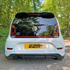 Puede incluir: Volkswagen Up! GTI blanco con parachoques trasero negro, luces traseras rojas y un alerón negro. El coche tiene una matrícula negra con el texto "EZM+" en ella. El coche está aparcado en una carretera asfaltada.
