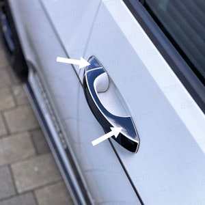 May include: A black and silver car door handle with a chrome accent. The handle is on a white car door.