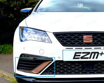 Puede incluir: Vista frontal de un coche blanco con una rejilla negra de panal y un logotipo de SEAT de color cobre. Un contorno rectangular azul resalta la toma de aire inferior izquierda, que contiene una pieza de acento de color cobre. El texto EZM+ es visible.