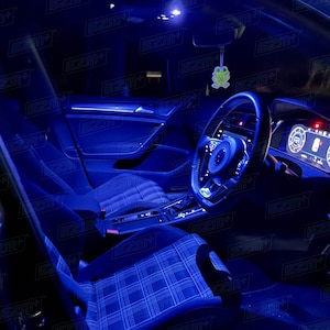 Puede incluir: El interior de un coche con luces LED azules iluminando el salpicadero, el volante y los asientos. Los asientos están cubiertos de una tela a cuadros azul y negra. Un pequeño ambientador de aire en forma de rana verde cuelga del espejo retrovisor.