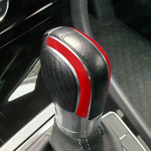 May include: Close-up of a black leather gear shift knob with a red stripe and silver accents. The knob has a textured surface and the word "DSG" is visible on the top.