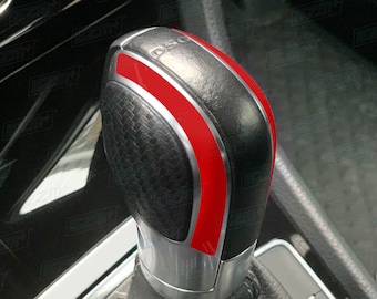 May include: Close-up of a black leather gear shift knob with a red stripe and silver accents. The knob has a textured surface and the word "DSG" is visible on the top.