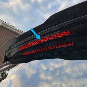 May include: Black car rear window with a red honeycomb-shaped taillight. The taillight is located on the top of the rear window and is surrounded by a black trim.