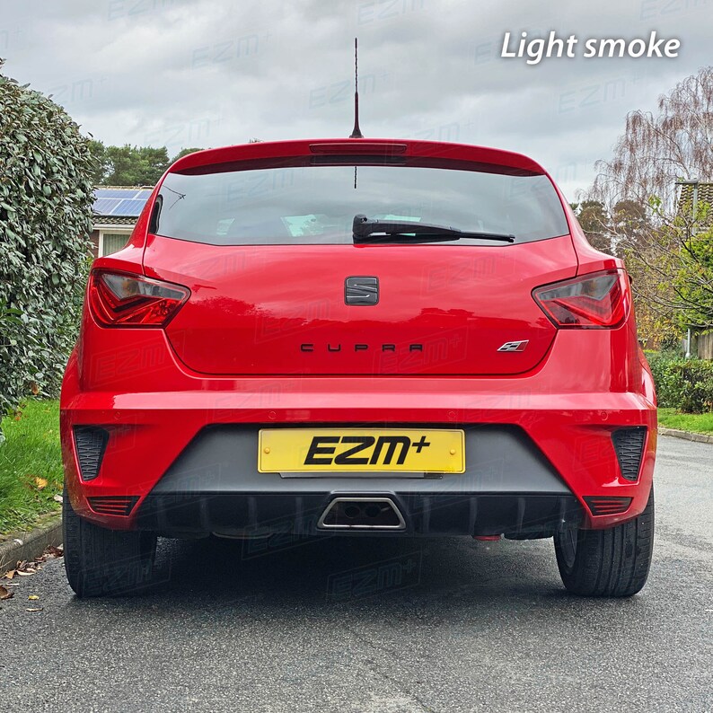 May include: Rear view of a red car with a black "CUPRA" emblem on the back. The car has a black rear bumper with a yellow license plate that reads "EZM+". The taillights are tinted a light smoke color.