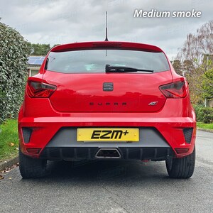 May include: Rear view of a red car with black tinted taillights and a black "CUPRA" emblem on the back. The car has a black rear bumper with a black exhaust pipe and a yellow license plate that reads "EZM+".