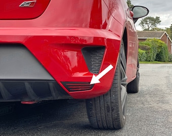 May include: Rear view of a red car with a black plastic bumper and a small red reflector on the lower right side. The reflector is outlined in black and has a series of horizontal lines.