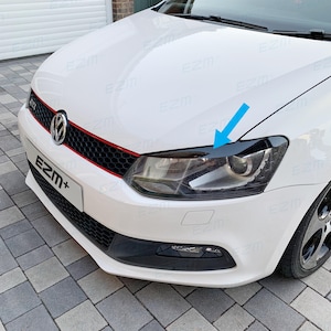 Op de afbeelding: Een witte Volkswagen Polo met een rode trim op de voorbumper. De auto heeft een zwarte voorbumper en zwarte koplampen. De koplampen hebben een zwarte trim rond de randen. De auto staat geparkeerd op een grijze stoep van klinkers.