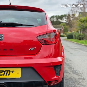 May include: Rear view of a red car with a black license plate that says "ZM+". The car has a black rear bumper with a black diffuser and a black spoiler. The taillights are red and clear. The car is parked on a street with a light smoke tint on the windows.
