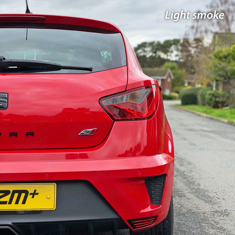 May include: Rear view of a red car with a black license plate that says "ZM+". The car has a black rear bumper with a black diffuser and a black spoiler. The taillights are red and clear. The car is parked on a street with a light smoke tint on the windows.