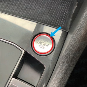 Op de afbeelding: Een close-up van een start-stopknop voor de motor van een auto. De knop is rond en heeft een rode ring eromheen. De woorden "Start Engine Stop" staan in wit op de knop.