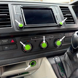 May include: A car dashboard with green-accented control knobs and buttons. The dashboard is black with a central display screen. The knobs are circular with a bright green centre. The image shows the interior of a vehicle.