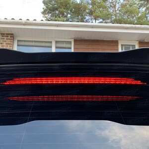Könnte beinhalten: Ein schwarzes Auto mit einem roten LED-Rücklicht. Das Rücklicht ist ein langer, dünner Streifen von Lichtern, der über die Rückseite des Autos verläuft.