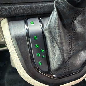 May include: Close-up of a car's gear shift lever. The lever is black with a silver trim and illuminated green letters indicating the gear positions: P, R, N, D, and S. The leather shift boot is black.