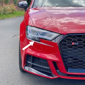 May include: A red Audi A3 with black accents on the front bumper and grille. The car has a black side mirror and a black headlight with a white arrow pointing to the headlight.