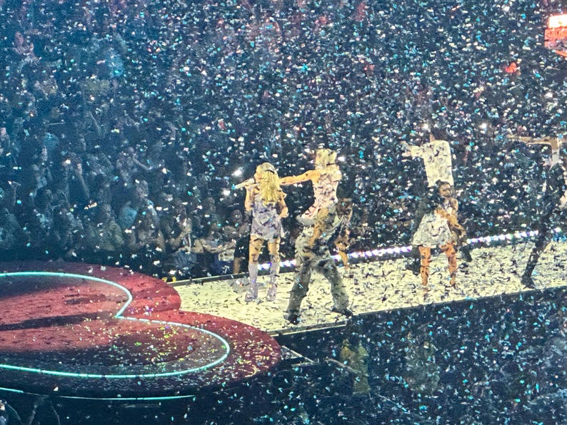 May include: A live concert stage with performers in colorful outfits, illuminated by stage lights and confetti. The stage has a heart-shaped design. The audience is visible in the background.