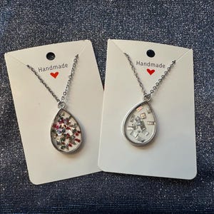 May include: Two silver-toned necklaces with teardrop pendants. One pendant contains red, green, and white accents, while the other has silver-colored square accents. Each necklace hangs on a silver chain and is displayed on a white card with the word "Handmade" and a red heart.