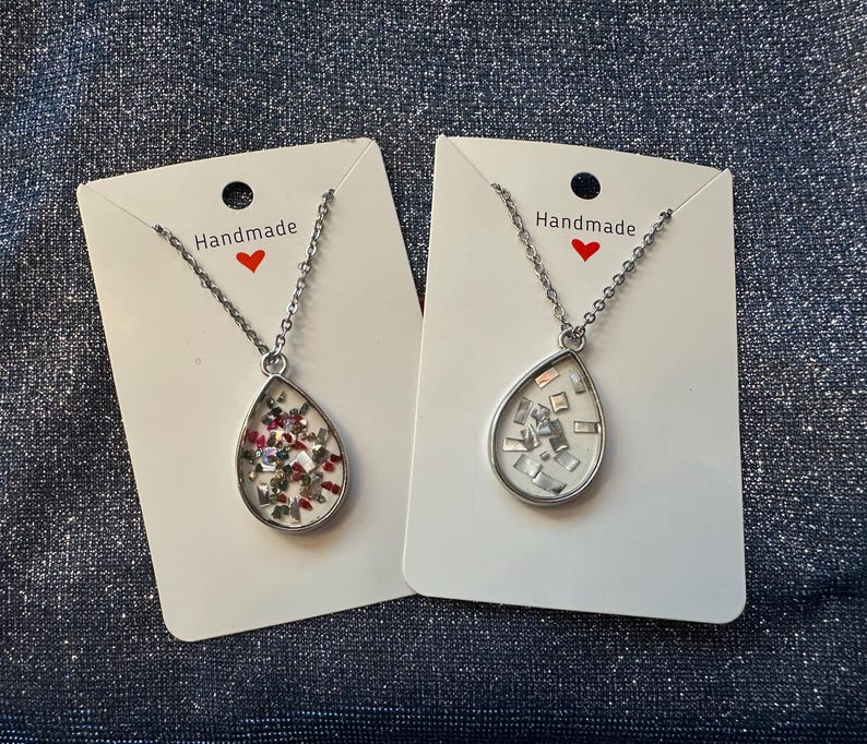 May include: Two silver-toned necklaces with teardrop pendants. One pendant contains red, green, and white accents, while the other has silver-colored square accents. Each necklace hangs on a silver chain and is displayed on a white card with the word "Handmade" and a red heart.