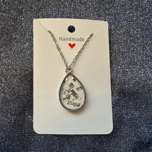 May include: A silver-toned necklace with a teardrop pendant. The pendant contains small, shiny, silver-colored rectangular pieces set against a white background. The necklace is displayed on a white card with the word "Handmade" and a red heart.