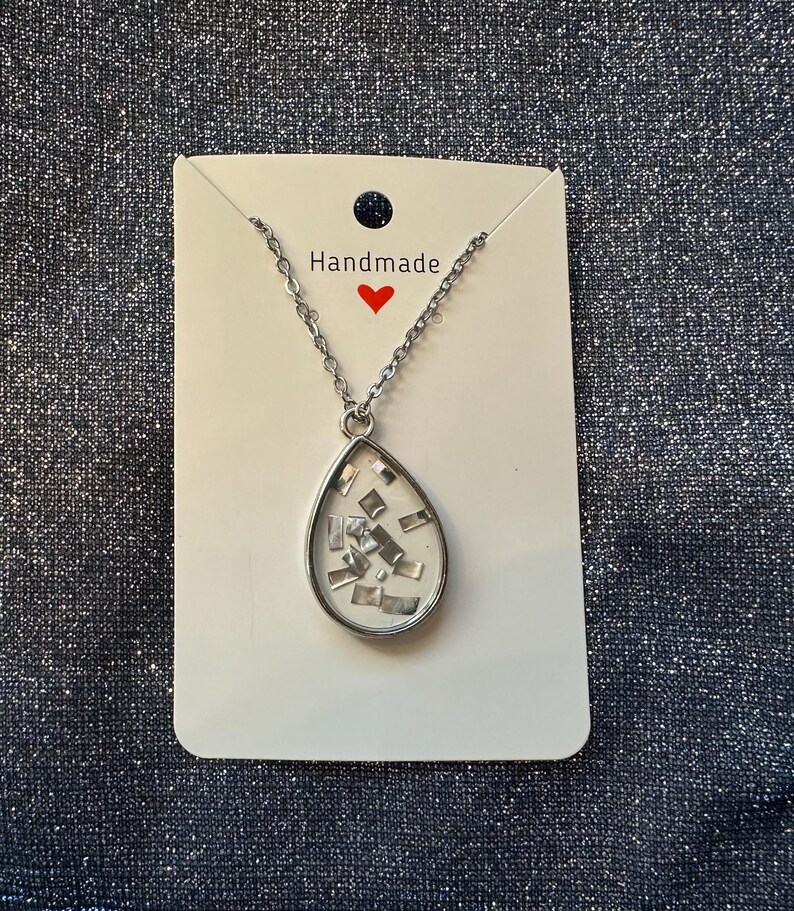 May include: A silver-toned necklace with a teardrop pendant. The pendant contains small, shiny, silver-colored rectangular pieces set against a white background. The necklace is displayed on a white card with the word "Handmade" and a red heart.