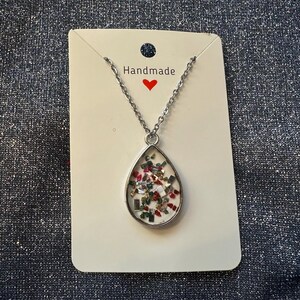 May include: Silver-toned necklace with a teardrop pendant filled with colorful confetti. The pendant hangs from a delicate chain and is displayed on a white card with the word "Handmade" and a red heart.