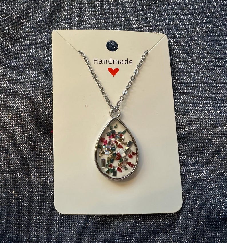 May include: Silver-toned necklace with a teardrop pendant filled with colorful confetti. The pendant hangs from a delicate chain and is displayed on a white card with the word "Handmade" and a red heart.