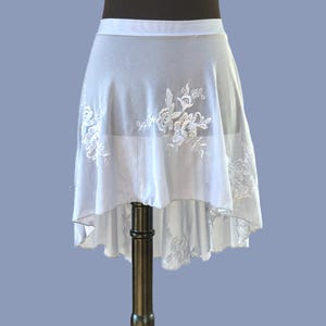 Könnte beinhalten: Ein weißer Ballettrock mit hohem und tiefem Saum. Der Rock hat zarte weiße Blumenstickereien auf der Vorder- und den Seiten. Der Stoff ist transparent und leicht, geeignet für Tanz oder Aufführungen.