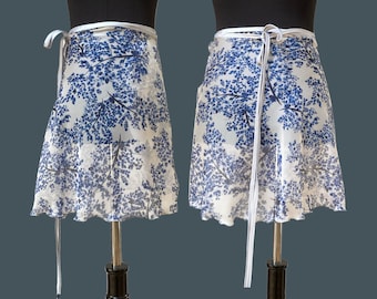 Op de afbeelding: Een wikkelrok met een witte achtergrond en een blauw bloemenpatroon. De rok heeft een witte striksluiting en een geplooid zoom. De rok wordt op een mannequin getoond.