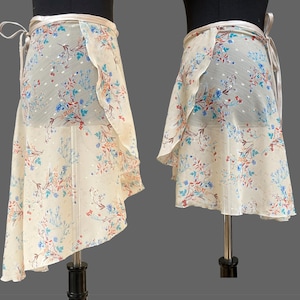 Pode incluir: Uma saia envelope em tecido transparente cor de creme com um delicado padrão floral em tons de azul, vermelho e castanho. A saia apresenta uma bainha assimétrica, um cós de cetim liso e longas fitas laterais para um fecho seguro.
