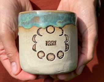 Könnte beinhalten: Eine Keramiktasse mit cremefarbenem Boden und türkisfarbenem Rand. Die Tasse zeigt ein geprägtes Design der Mondphasen und die Worte "MOON PHASES". Die Tasse wird in zwei Händen gehalten.