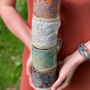 Può includere: Una pila di quattro tazze in ceramica con un motivo a fiore di loto. Le tazze sono in tonalità di marrone, bianco, verde e blu.