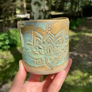 Op de afbeelding: Een keramische mok met een lichtblauwe glazuur en een bruine rand. De mok heeft een reliëfdesign van een mandala met een lotusbloem in het midden.