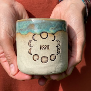 Könnte beinhalten: Eine Keramiktasse mit cremefarbenem Boden und türkisfarbenem Rand. Die Tasse zeigt ein geprägtes Design der Mondphasen und die Worte "MOON PHASES". Die Tasse wird in zwei Händen gehalten.