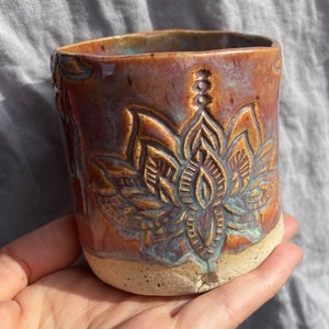 Könnte beinhalten: Eine handgefertigte Keramiktasse mit einem Lotusblumen-Design. Die Tasse hat eine braune und blaue Glasur mit strukturierter Oberfläche. Der Boden der Tasse ist unglasiert und zeigt die natürliche Tonfarbe.