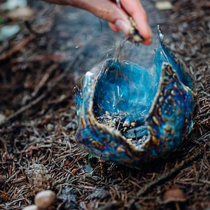 May include: A blue and gold ceramic bowl with a crackled glaze sits on a bed of pine needles. Smoke rises from the bowl, which contains burning incense.